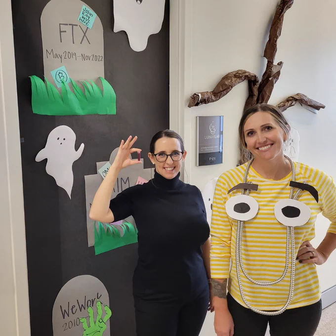 Lunarlab co-founders Elizabeth Anderson (on the left) and Kelli Lucas (on the right), standing in front of an office door adorned with a graveyard-themed decoration. Elizabeth is seen holding a vial of "red substance" (the red substance is not real).