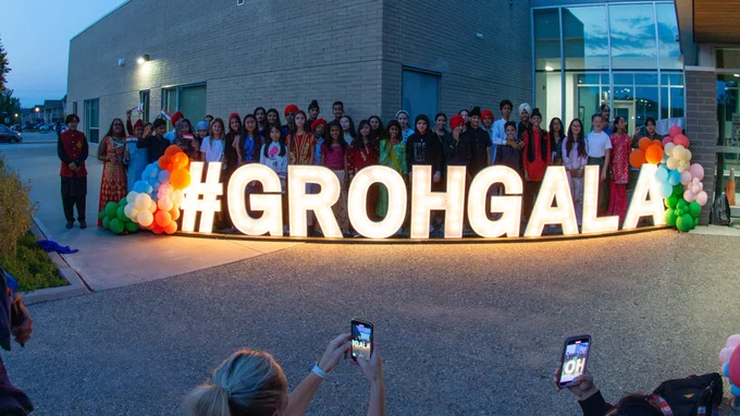 A group of students standing behind a lit up sign that says #GrohGala. They are in front of a school.