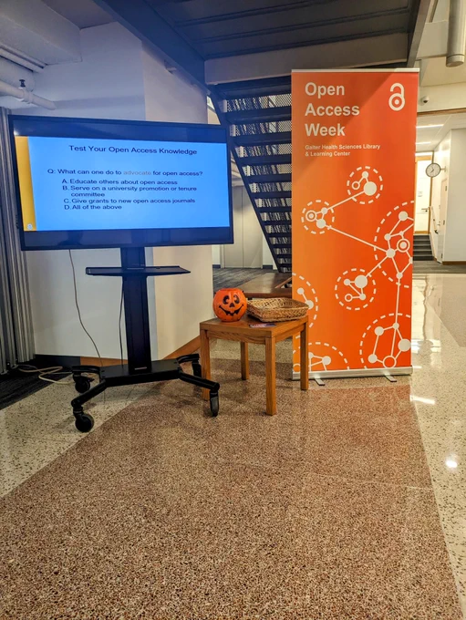 Library atrium with a digital monitor showing an open access themed powerpoint beside a side table with a jackolantern and a standing banner showcasing the open access logo