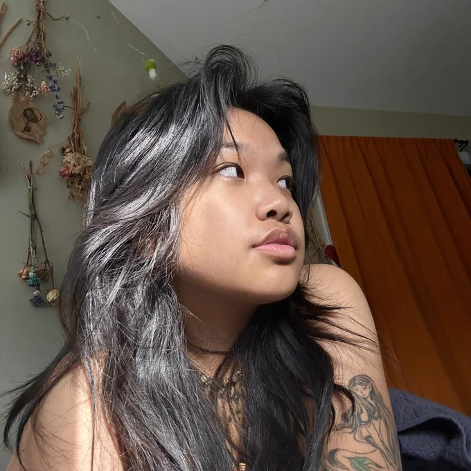 anata laylay looking off to the side with the sun highlighting their hair. their sirena tattoo on their arm is showing. they have dried flowers and an orange curtain in the background.