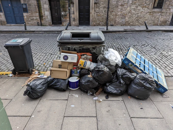 A pile of bags, boxes and other commercial waste around a street side wheelie bin.