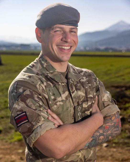 Captain Richard Ainsworth-Masiello ARCC stands smiling wearing his Army uniform, a dark grey beret and his arms folded. In the background there are fields and on the horizon a mountain range.