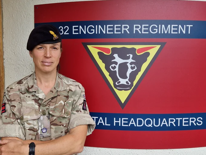 Major Alanda Scott KCVS stands arms folded wearing an Army uniform and a dark beret. Behind her there is a red flag with the words '32 Engineer Regiment' an emblem of a bull and  'Headquarters'. 