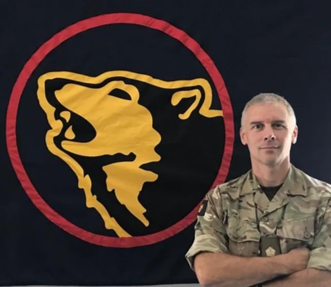Brigadier Chris King CBE stands with his arms folded in Army uniform in front of an emblem representing a wolf within a circle. 