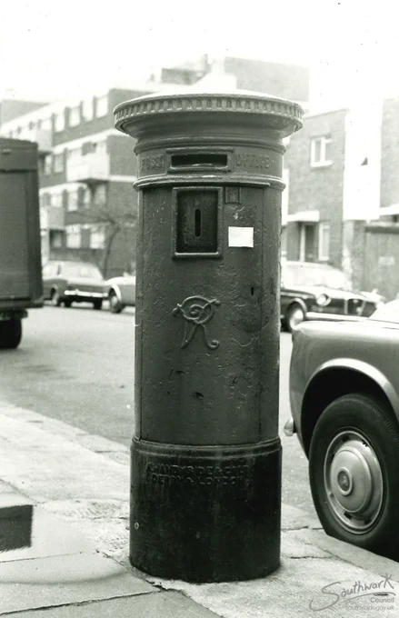 Black and white photograph showing a Victorian post box with an engraving 'VR' on the front. The post box is standing on a pavement on Lomond Grove, Camberwell, London. A car's wheel is shown next to the post box frame right and there are flat and cars in the background.