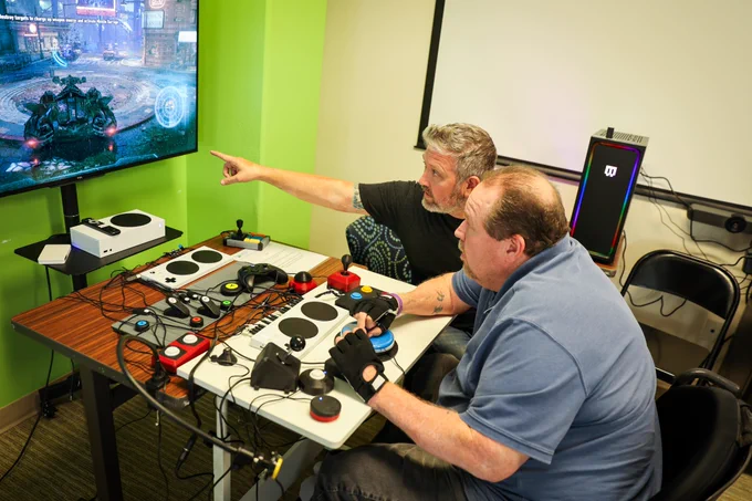 Erik instructing a client sitting in a wheelchair with an adaptive gaming setup. He's playing a Batman game on the xbox