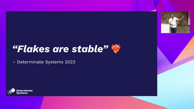 A slide with a dark purple background, and the text has a quote in italics bold "Flakes are stable" with a heart-on-fire emoji. The quote is attributed to Determinate Systems, 2023, since the upstream project has not yet committed to stable Flakes -- but we have. In the corner of the screen is @grhmc on stage, presenting on the benefits of flakes and why they are the future of Nix.