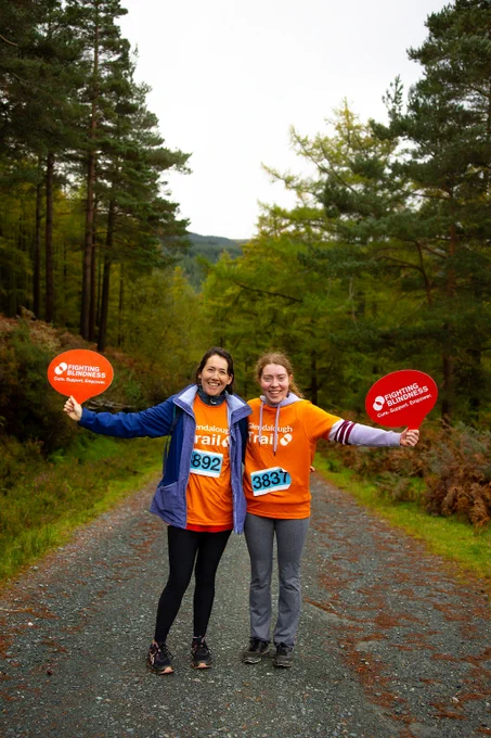 Tess McCann and Grace Ruddin posing with two Fighting Blindness speech bubble boards with the Fighting Blindness logo on them. They are in the middle of the Glendalough trail run.