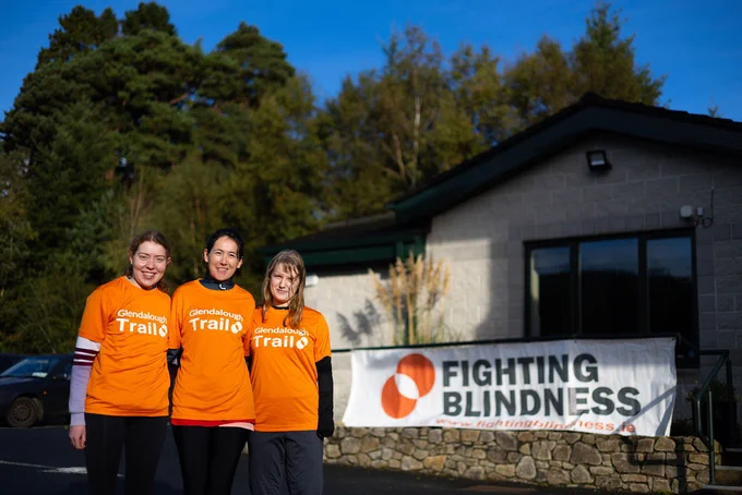 The image shows Grace Ruddin, Elin Strachan and Tess McCann smiling in front of a Fighting Blindness banner, wearing their Glendalough Trail Run branded t-shirts.  