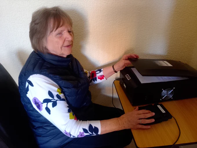Margaret is sat at a table using her ReadEasy reading machine.