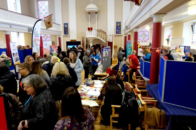 A busy indoor venue being used as an information market place, full of tables spread with leaflets, display stands and people from different groups and services which support children and families in Moray, talking to ones another.