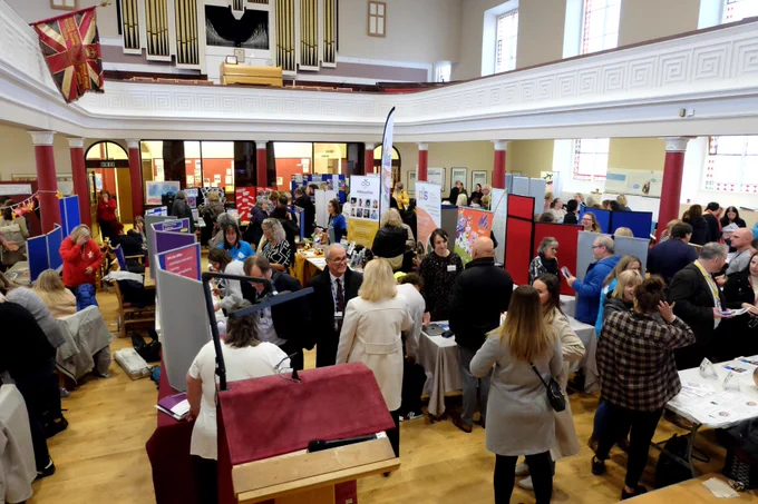 A slightly different view of the busy indoor venue being used as an information market place, full of tables spread with leaflets, display stands and people from different groups and services which support children and families in Moray, talking to ones another.