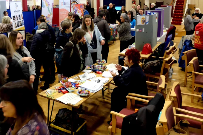 A slightly different view of the busy indoor venue being used as an information market place, full of tables spread with leaflets, display stands and people from different groups and services which support children and families in Moray, talking to ones another.