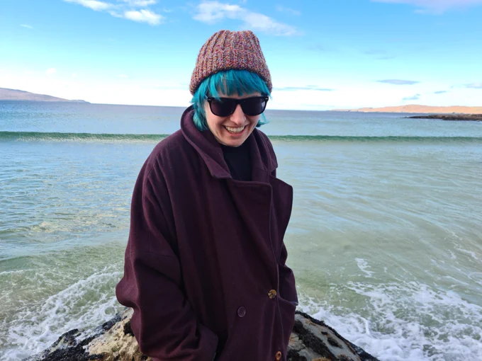 A woman in a coat, wooly hat and sunglasses, standing on a rock next to the sea. The sky is blue and the water is calm.