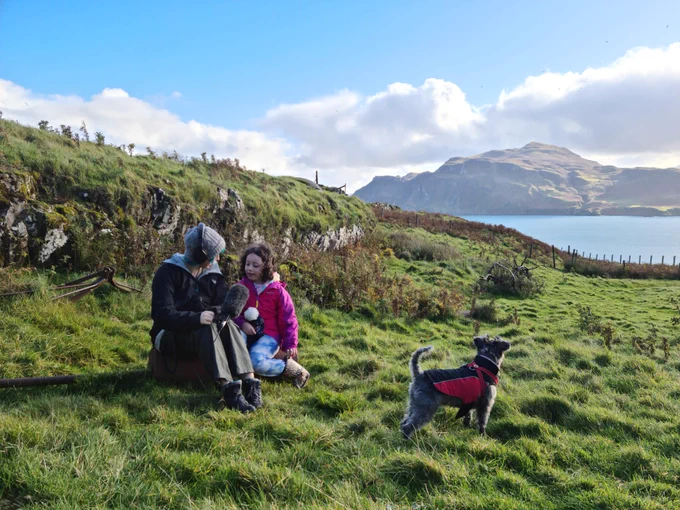 A woman interviewing a young girl outdoors, on a hillside, near a body of water, with a small dog nearby.