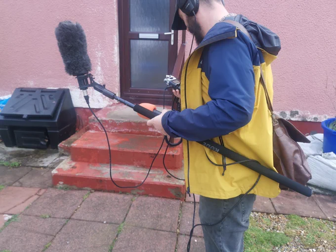 A man with a boom mic, recording device and headphones, standing next to the door of a pink building.