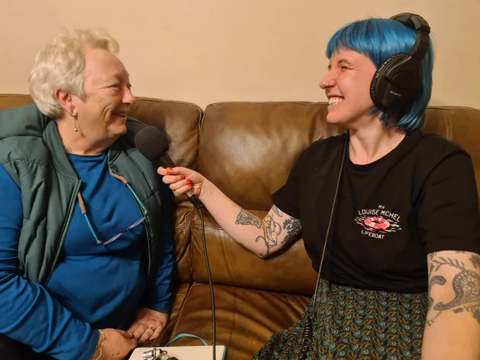 One woman interviewing another woman, holding up a microphone and wearing headphones. Both are sitting on a brown leather sofa.