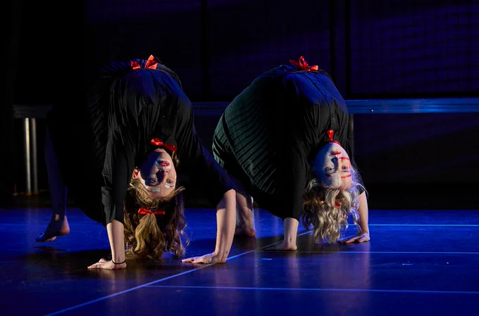@unforgottengen perform The Gift. Two performers dressed in black with black, white, and red painted faces, and red hair bows, crab walk backwards across the stage.
