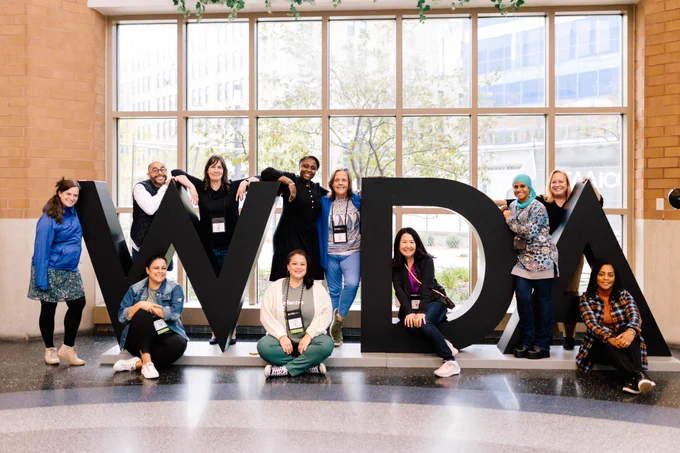 Eleven people gathered around giant letters that spell out "WIDA." The "I" is missing.