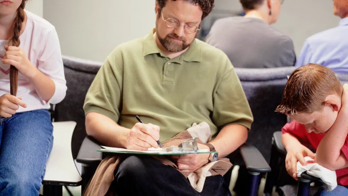 A man is sitting in a waiting room filling out paperwork on a clipboard. He is surrounded by other people.