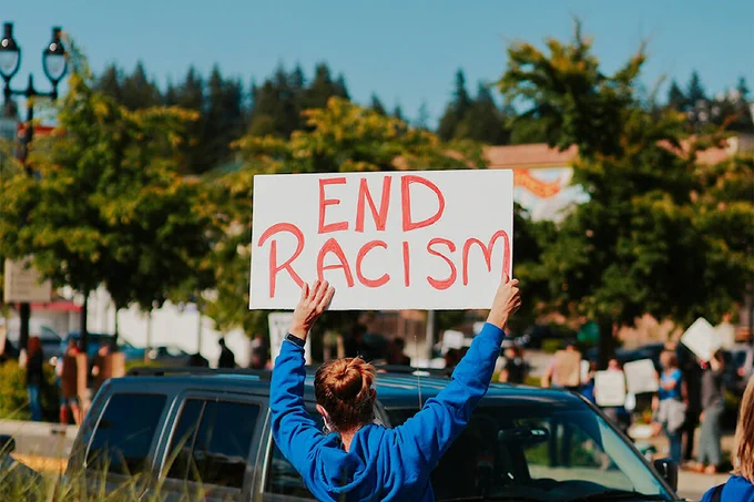 Person holds a protest sign over their head, red lettering on white poster board: "End Racism." Source: Unsplash.