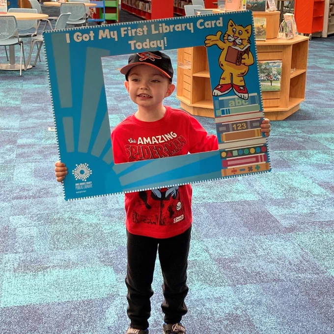 young boy in a red shirt and hat smiling and holding up a library cardboard cutout saying "I got my first library card today!"