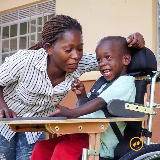 A five-year-old boy with his mother in Uganda. The boy is smiling in a wheelchair as his mother bends over him.