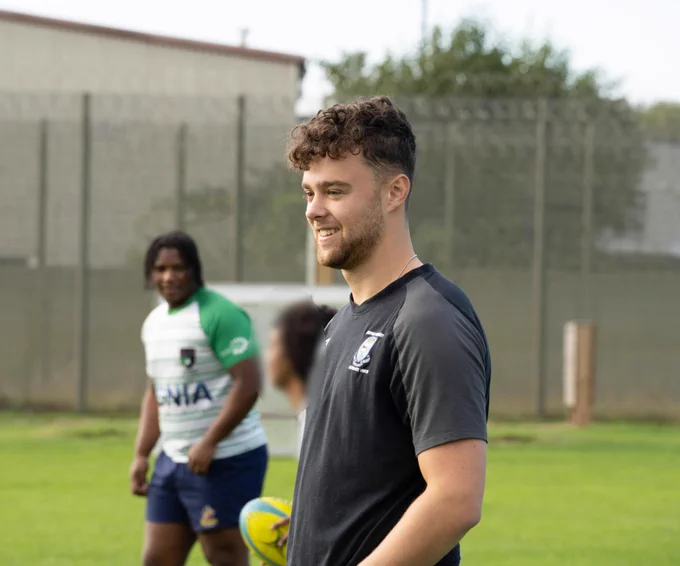 Photograph of a young rugby player smiling with two rugby players in the background. 