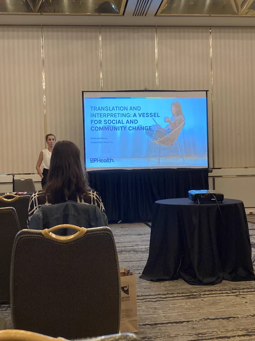 Photo of a presenter standing next to a PowerPoint slide that says “Translation and Interpreting: A Vessel for Social and Community Change”
