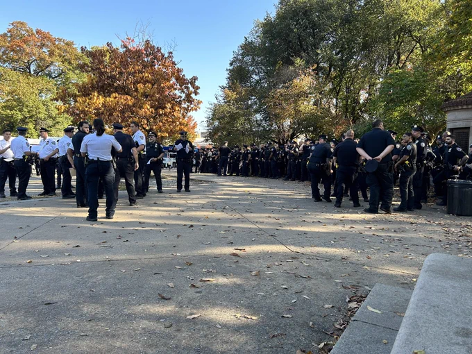 A long row of uniformed police officers stands on a wide sidewalk, with another row fanning out to the left of them.