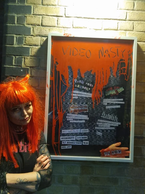 Woman with long red hair fashioned into devil horns in front of event poster with red paint and cut out type on black background, on a wall at night with overhead lighting 