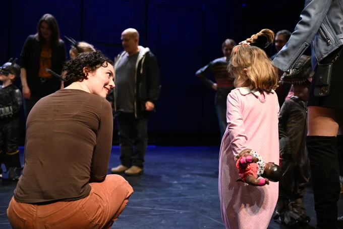 A group of people stand in a circle on a stage. In the foreground, a woman engages with a small child in a long, pink gown.