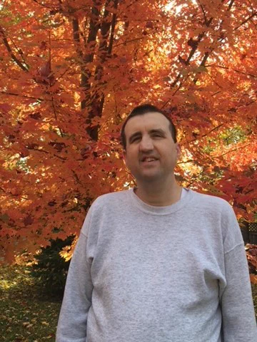John wears a grey shirt and smiles at the camera in front of a tree with orange leaves.