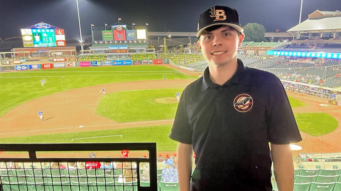 Max Oxley, wearing a team hat and polo shirt, poses for a photo in front of the Louisville Bats' field.
