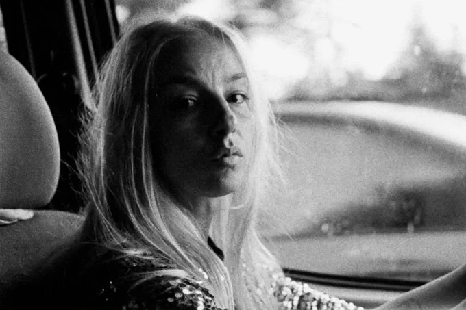 Black and white image of a person with blond hair and light-toned skin looking at us as they drive a car
