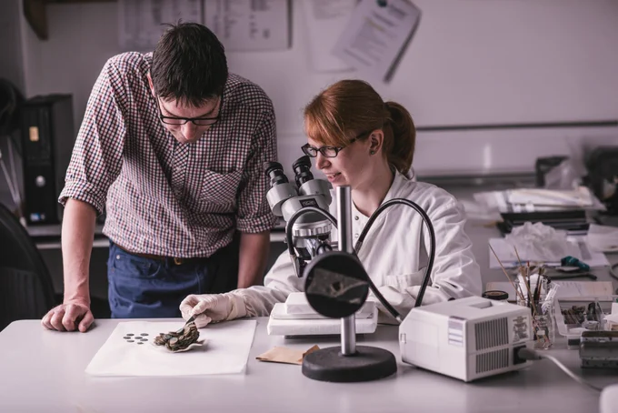 A woman in a white coat is seated at a microscope. She is wearing gloves and handling a small selection of coins, showing them to a man standing next to her. They are in a lab setting.