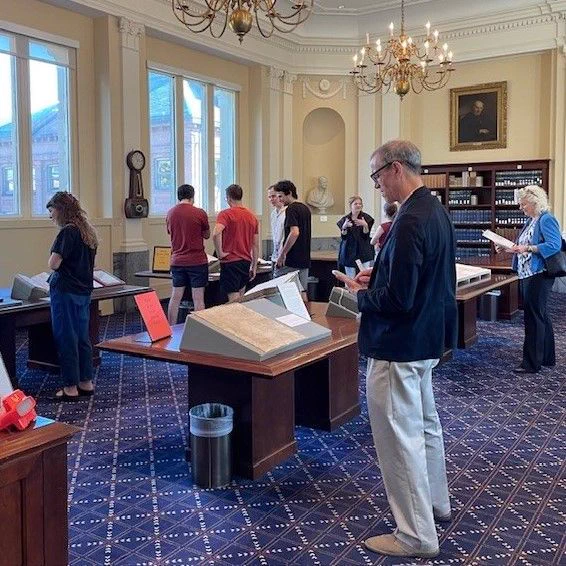 People stand around four tables in a room with yellow walls, a chandelier, and a blue patterned carpet, looking at materials displayed on the tables. 