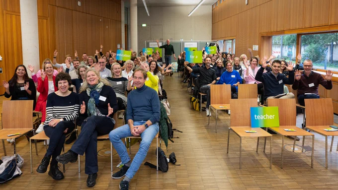 A cheerful group of attendees in a well-lit auditorium, seated and waving at the camera. Many participants are smiling and seem engaged. In the foreground, a few individuals are sitting on wooden chairs, with a sign labelled 'teacher' placed on an unoccupied seat. In the background, banners displaying words like 'education' and 'future' are visible.