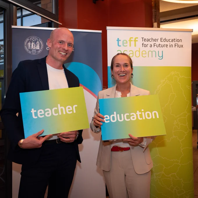 Two cheerful individuals, a man and a woman, standing in front of promotional banners for 'Teacher Education for a Future in Flux' academy. The man, wearing a dark jacket and white shirt, holds a sign reading 'teacher'. Beside him, the woman, dressed in a light-coloured suit, holds a sign reading 'education'. The atmosphere is positive, and the venue has a modern interior with hints of a catering area in the background.