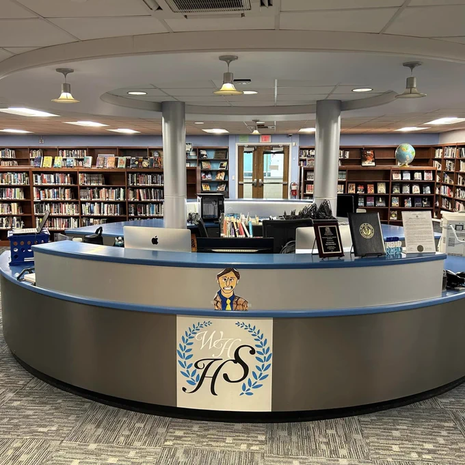 A blue and grey circulation desk of a library with the Woodland Hills High School logo on it. Library bookshelf stacks can be seen in the background. 