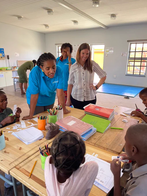 Three women standing at a classroom desk looking on  where a group of four children sits around their classroom desk doing arts and crafts 