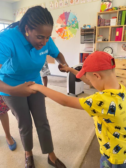 Woman with cyan color t-shirt holding hands with a young boy child with learning disability, wearing a yellow shirt with a red cap on his head, to support him with taking a few steps