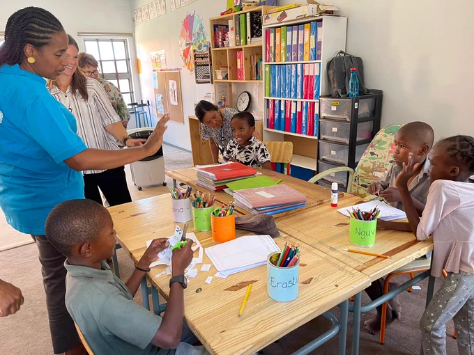 Two women standing at a classroom desk looking on while a group of five children sit around the desk showing their arts and crafts 