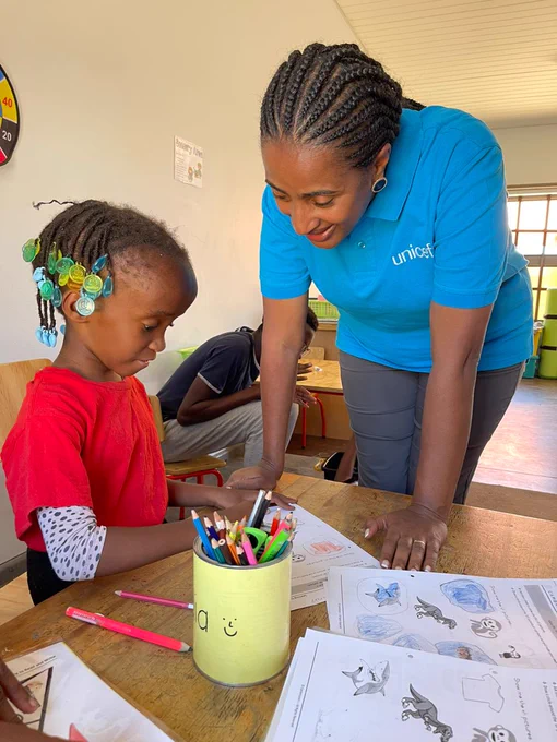 Woman in cyan color t-shirt, watching on while young girl child with learning disability, dressed in red shirt shows off her coloring page