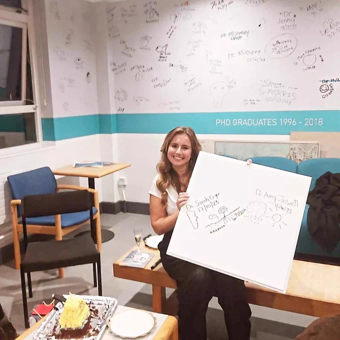 Sarah, a white woman, holds a ceiling tile with Dr Sarah Cryer 17/10/2023 written on it and a sketch of mangroves and seagrass. 