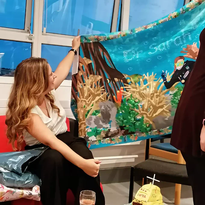 Sarah holds up a towel which is printed with mangroves and corals, bubbles spell out Dr Sarah 