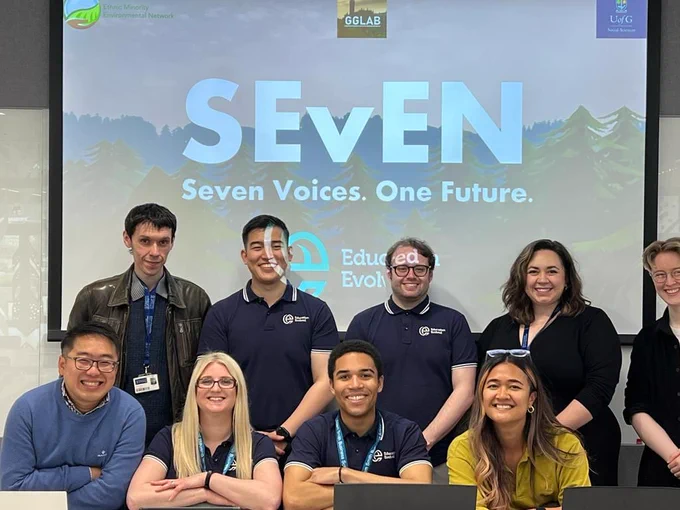 A group photo of the team behind the creation of the video game. Behind them is a screen which reads: Seven - Seven voices, one future. 