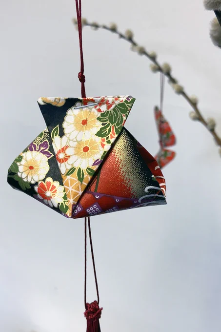 a hanging origami decoration in the shape of a Chinese money bag is visible in the foreground with an origami butterfly decoration visible in the background hanging on a pussy willow branch. 