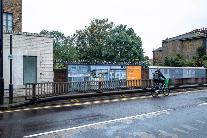 Picture 2 is of a billboard on street behind railing. Billboard advertises At Home in Hackney exhibition with 3 photos of closed down shopfronts. 