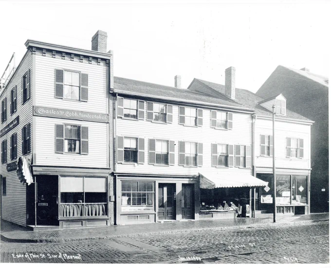 Cobblestone street with wooden frame storefronts including Photograph shows: Charles W. Cobb Undertaker, R. J. Tash Successor to Mumler's 